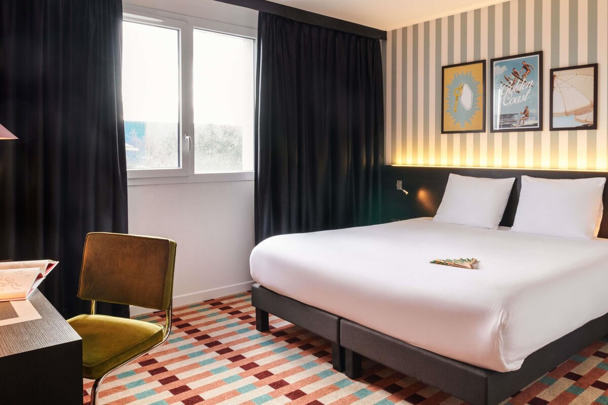 A hotel room with a bed, a desk, a chair, a window with curtains, and wall art.