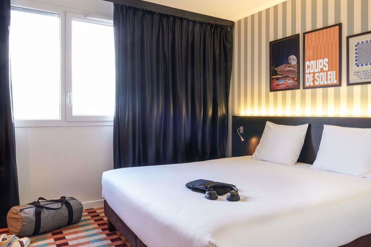 A hotel room with a bed, a window with curtains, a wall with striped wallpaper, and a poster.