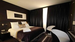 Habitación de hotel con una cama grande, una mesita de noche, una silla y un ventanal con cortinas.