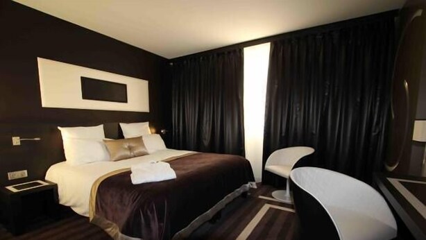 Habitación de hotel con una cama grande, una mesita de noche, una silla y un ventanal con cortinas.