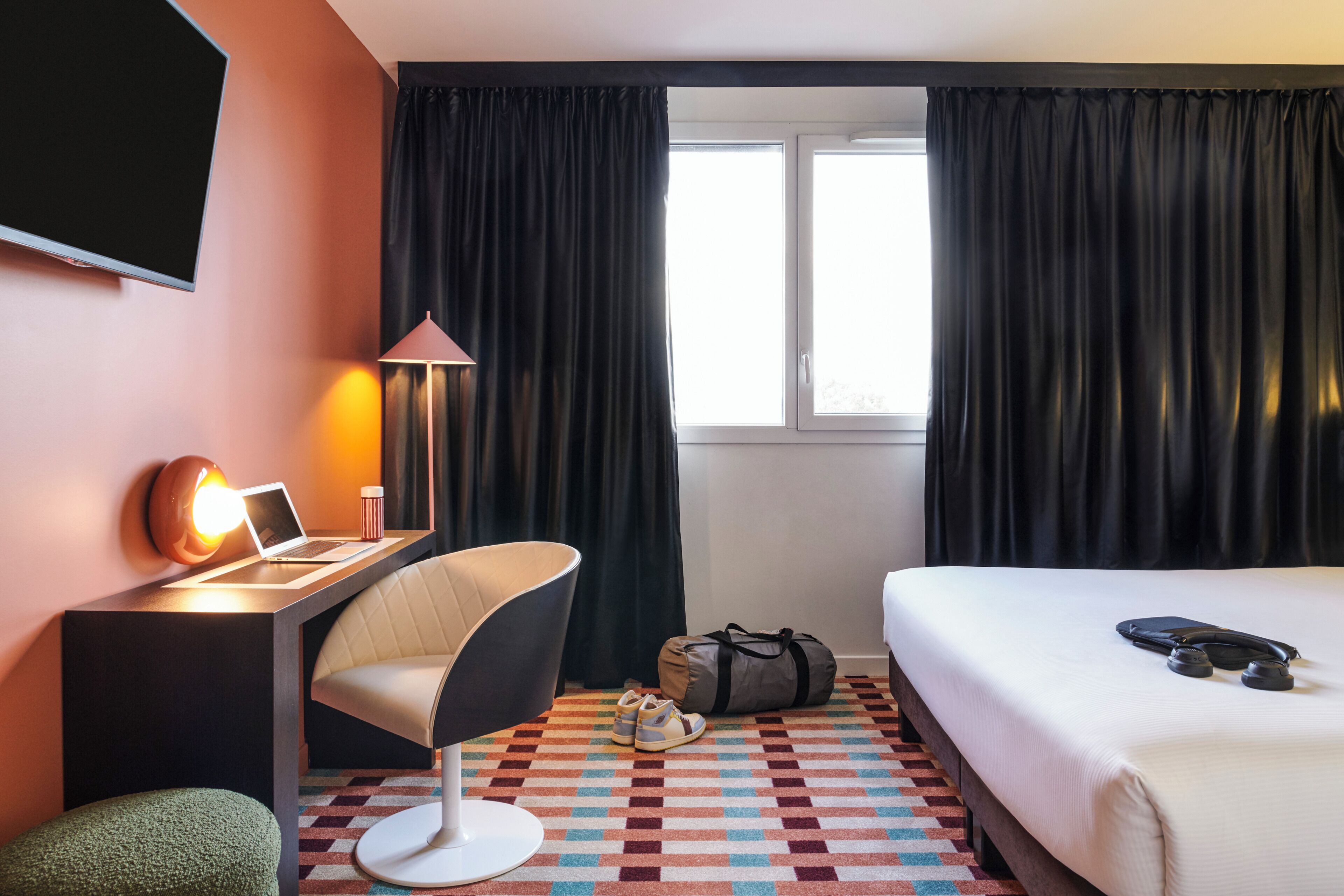 Habitación de hotel con cama, escritorio con computadora portátil, silla, lámpara, televisor y ventana con cortinas.