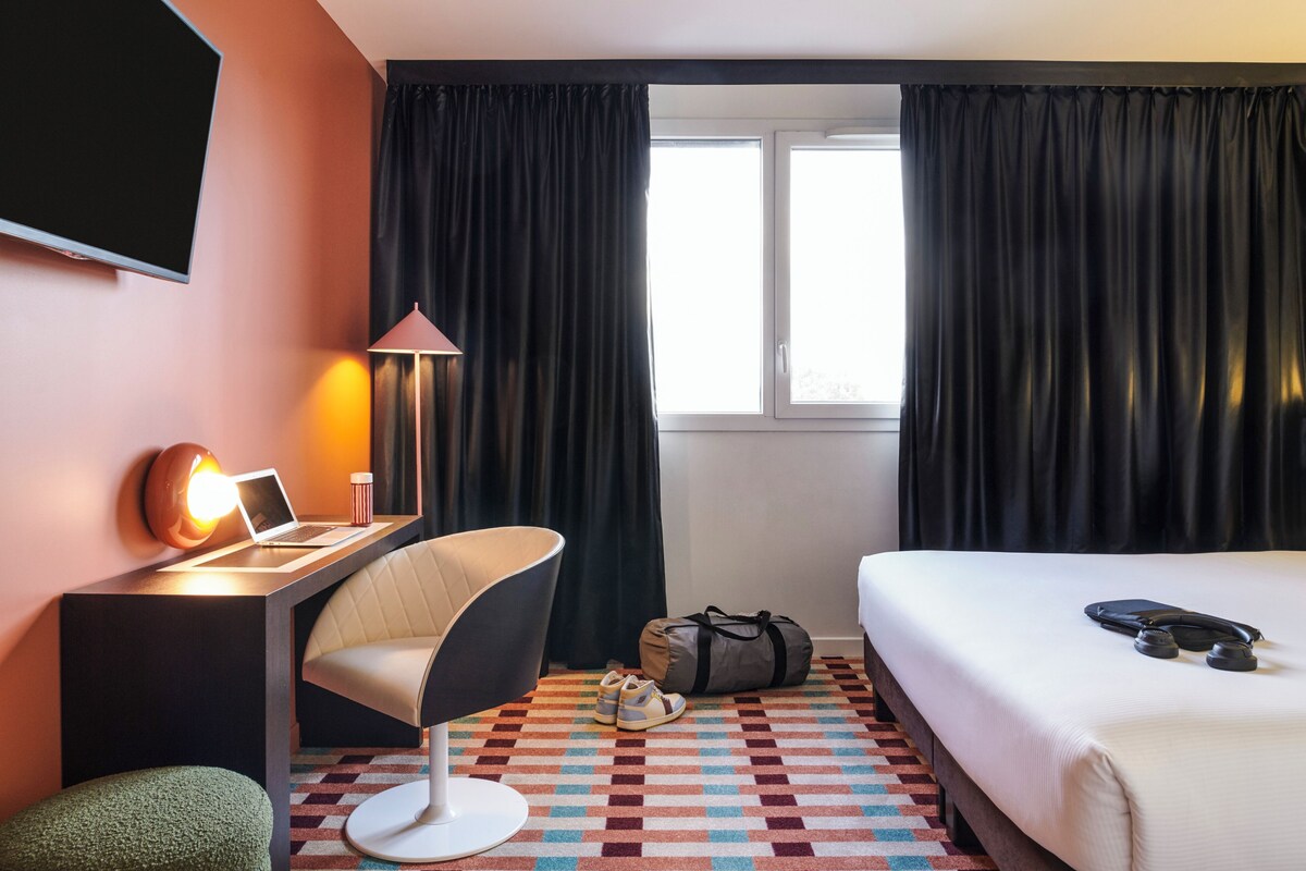 Habitación de hotel con cama, escritorio con computadora portátil, silla, lámpara, televisor y ventana con cortinas.