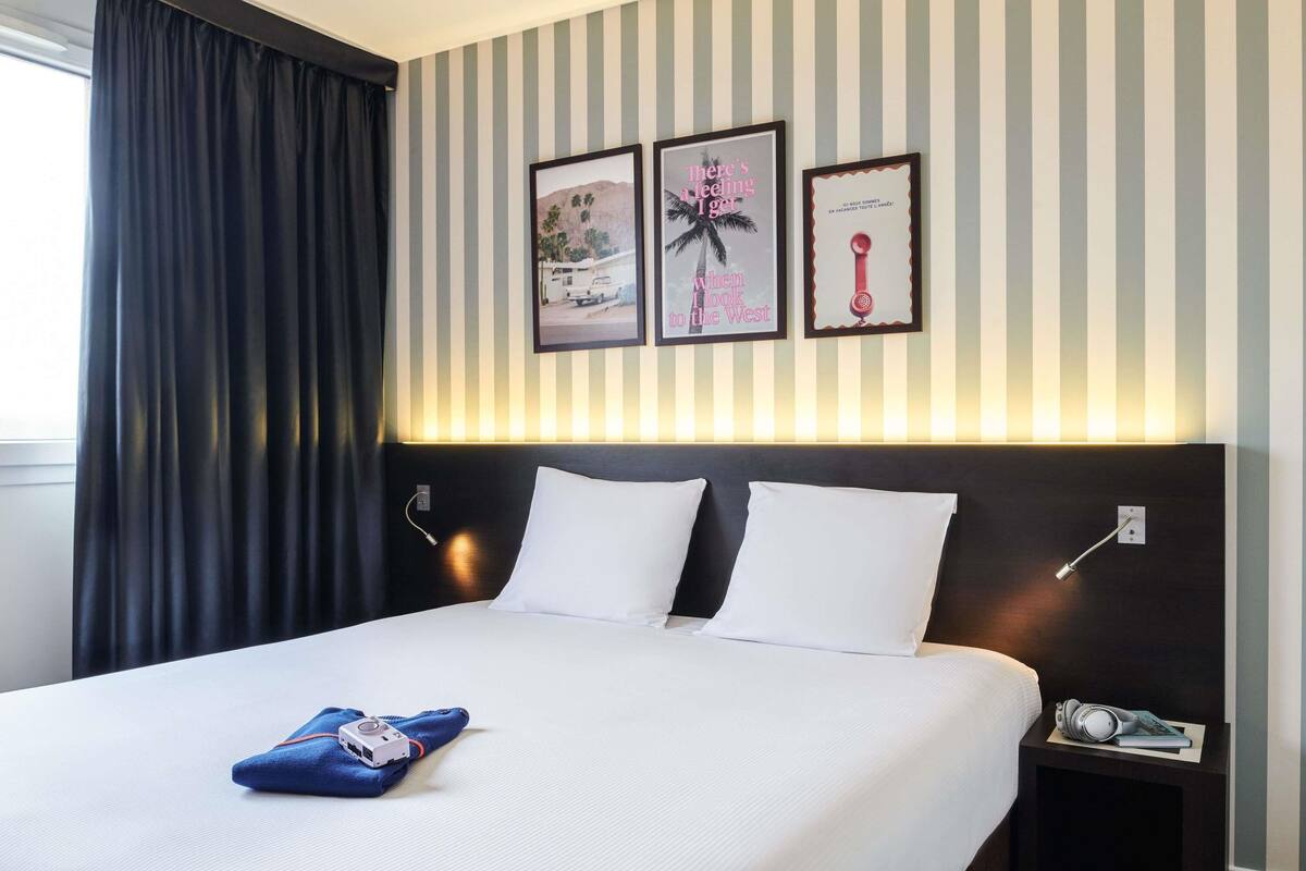 Una habitación de hotel con cama, mesita de noche y pared con papel tapiz a rayas y cuadros enmarcados.