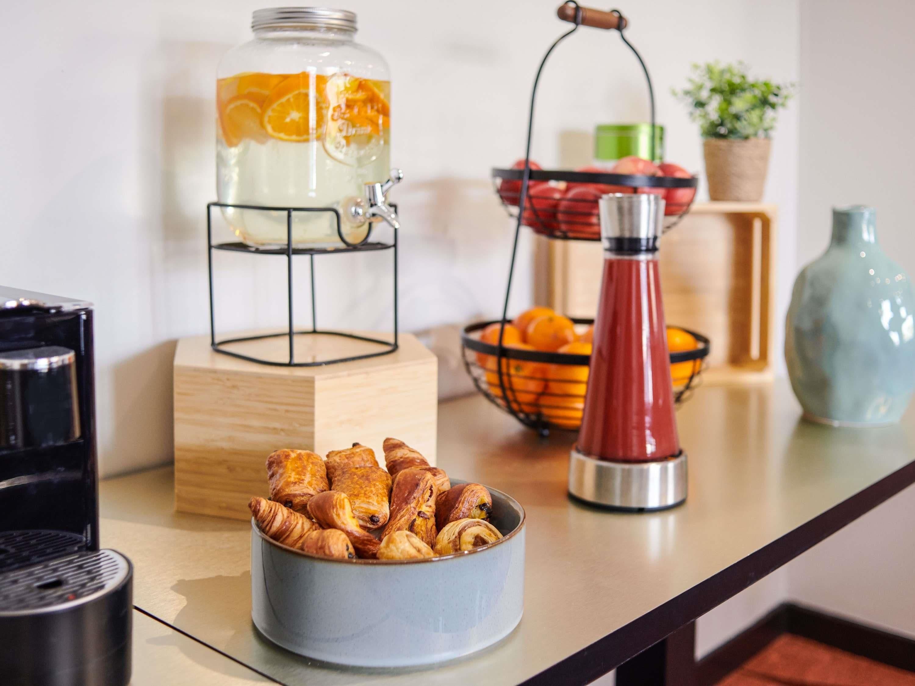 A breakfast setup with a fruit-infused water dispenser, a bowl of pastries, a fruit basket, and a coffee machine.