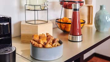 A breakfast setup with a fruit-infused water dispenser, a bowl of pastries, a fruit basket, and a coffee machine.