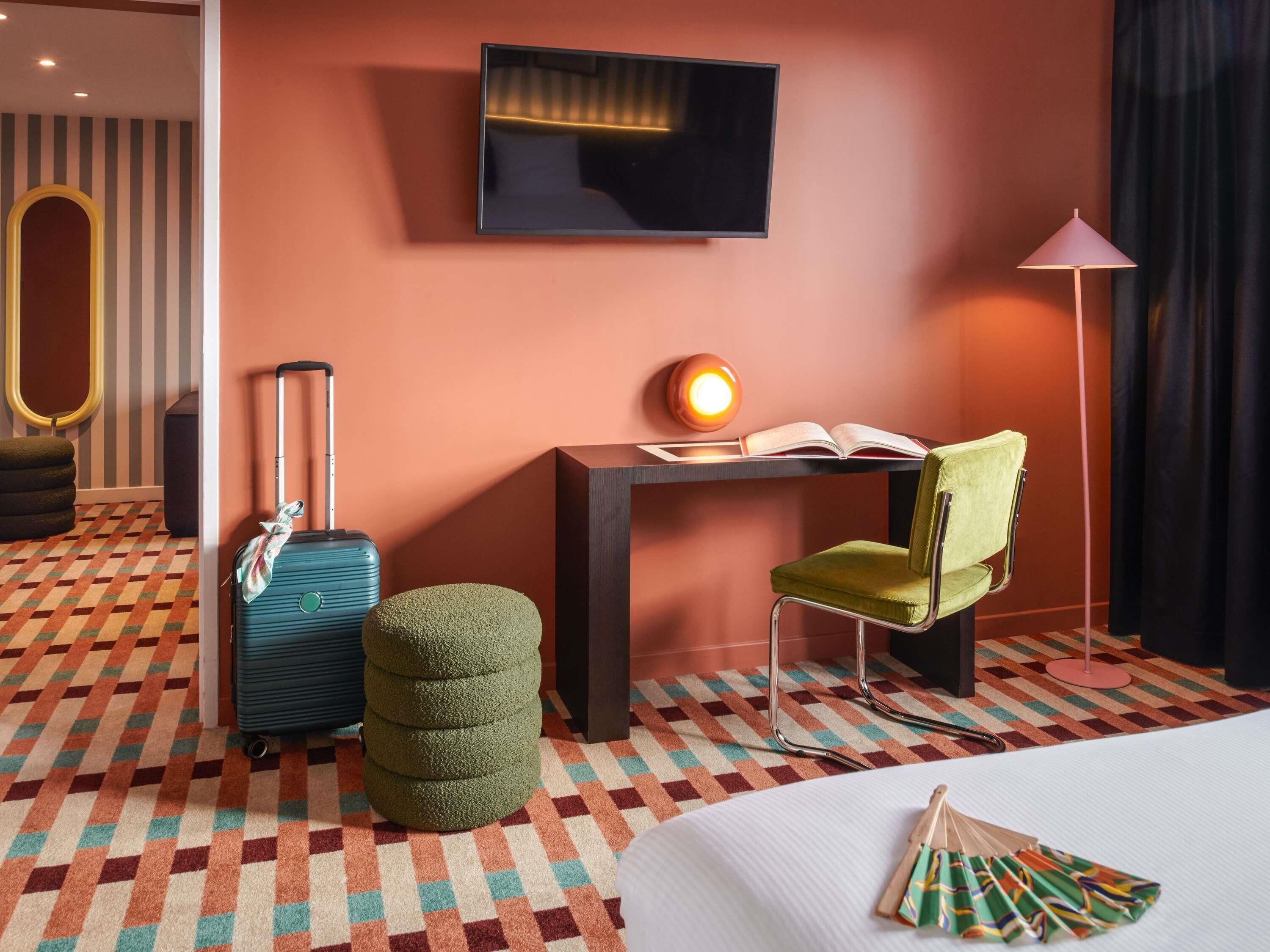 A hotel room with a bed, a desk with a lamp, a chair, a suitcase, and a small ottoman.