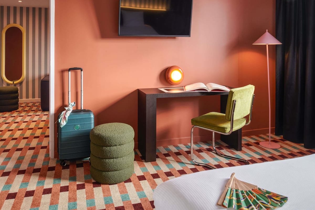 Habitación de hotel con una cama, un escritorio con lámpara, una silla, una maleta y un pequeño reposapiés.