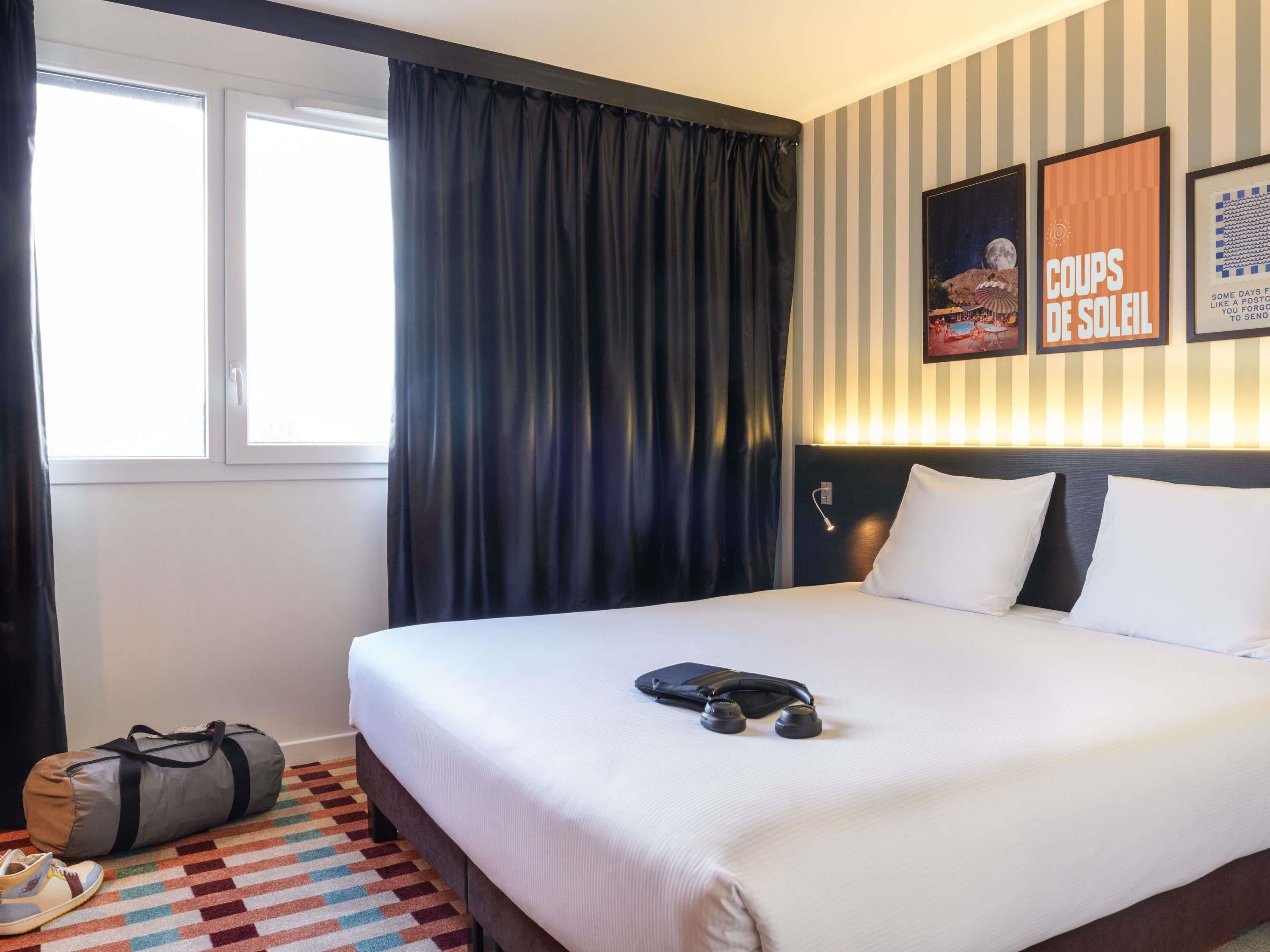 Une chambre d’hôtel avec un lit, une fenêtre avec des rideaux, un mur recouvert d’un papier peint à rayures et une affiche.