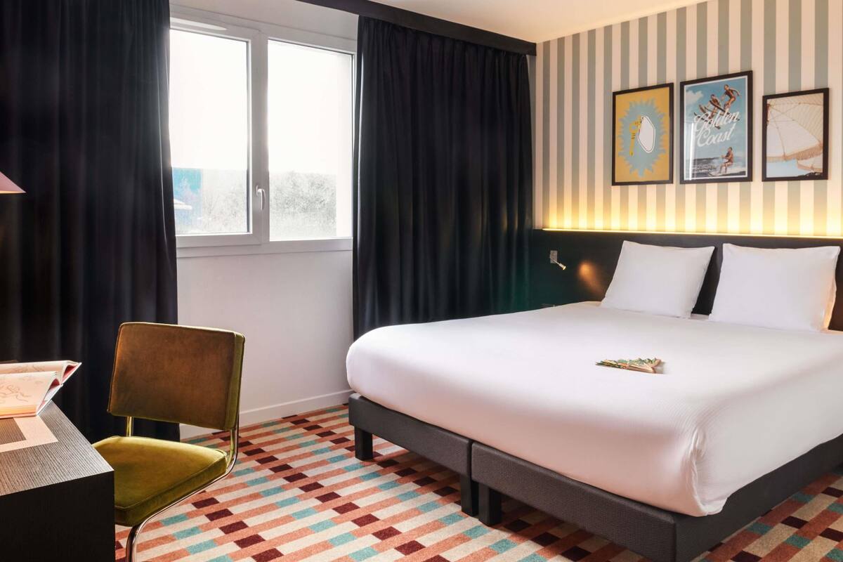Habitación de hotel con cama, escritorio, silla, ventana con cortinas y decoración en la pared.