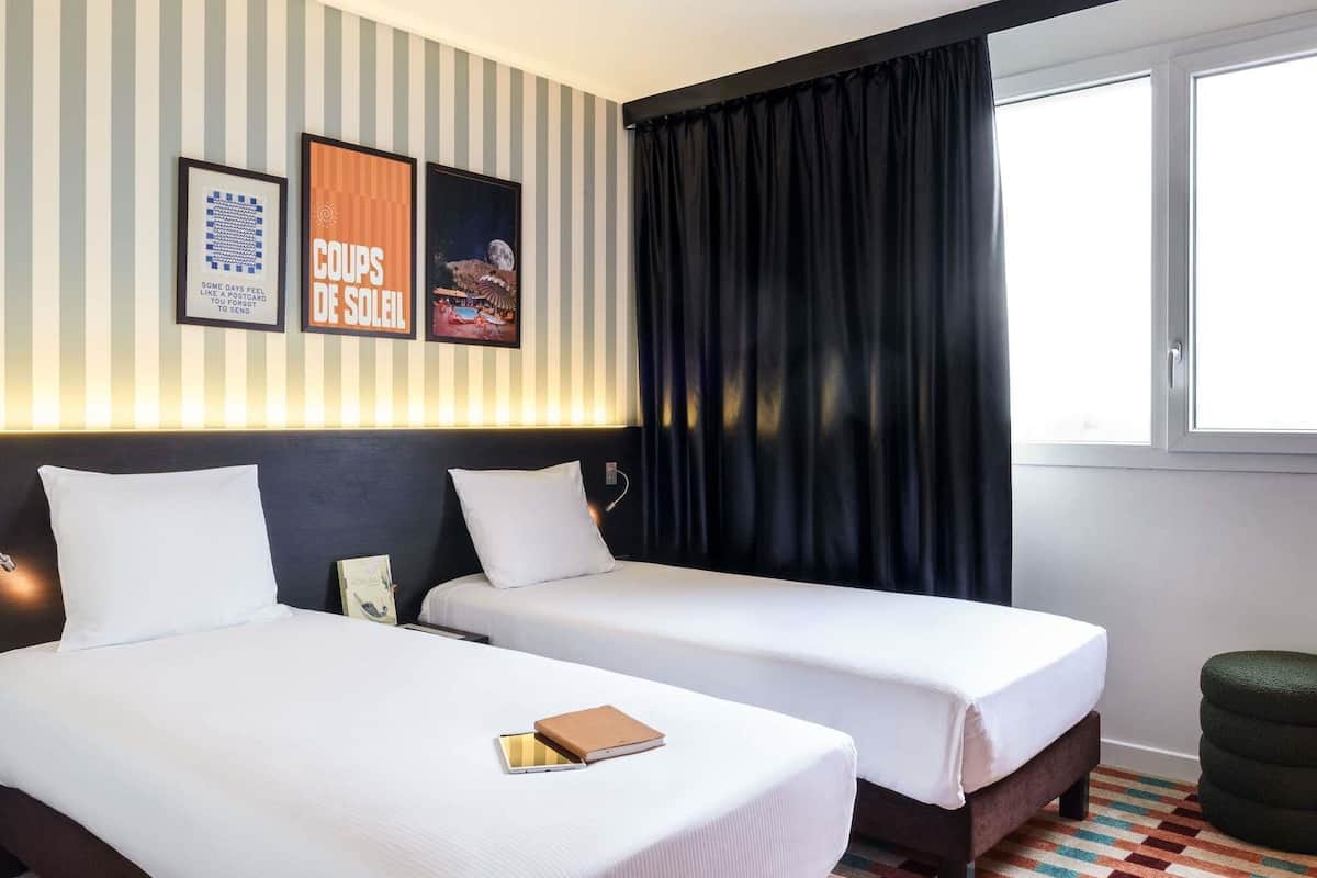 A hotel room with two beds, a window with curtains, striped wallpaper, and framed pictures on the wall.