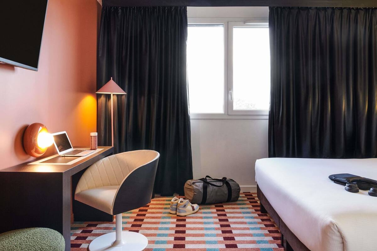A hotel room with a bed, a desk with a laptop, a chair, a lamp, a window with curtains, and a patterned carpet.