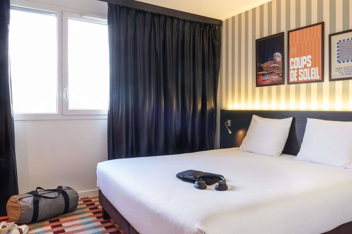 Una habitación de hotel con una cama, un ventanal con cortinas, una pared con papel tapiz a rayas y un cartel.