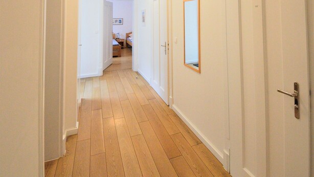 Un pasillo con suelo de madera, paredes blancas y una lámpara en el techo. Hay puertas cerradas a la derecha y una puerta abierta al final que da a una habitación con una cama y una mesita de noche.