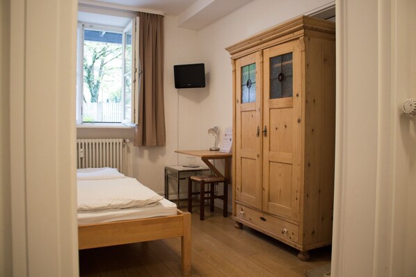 A bedroom with a wooden bed, a desk with a chair, a wardrobe, and a television on the wall.