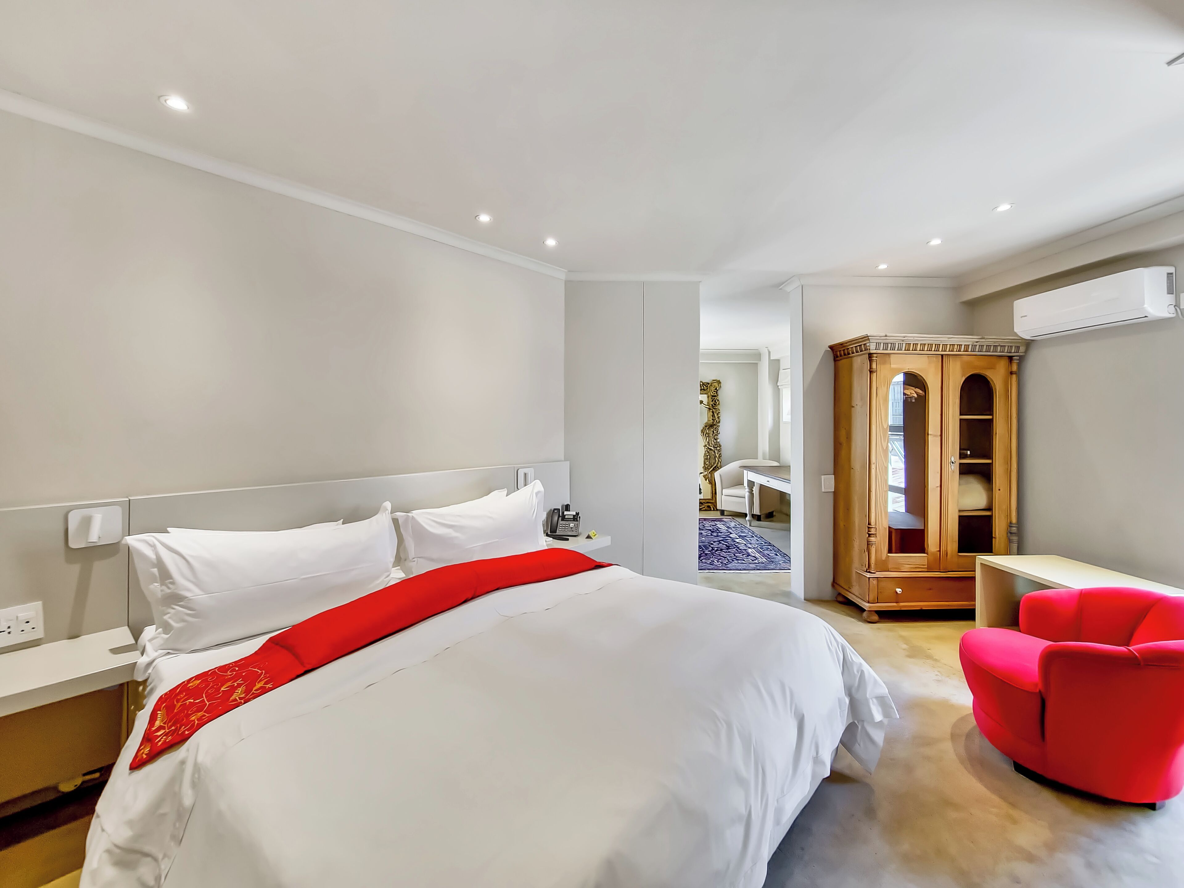 A hotel room with a bed, a red chair, a wardrobe, and an air conditioning unit.