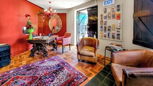 A room with a wooden floor, a red wall, a mirror, a chandelier, a table with a vase, a chair, and an exit sign. - Rouge on Rose Boutique Hotel (Cape Town)