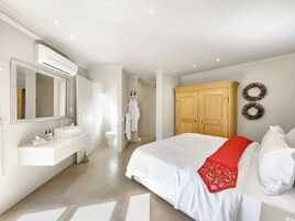 A modern bedroom with a large bed, a wooden wardrobe, a white bathroom with a sink and mirror, and a hallway leading to a shower.