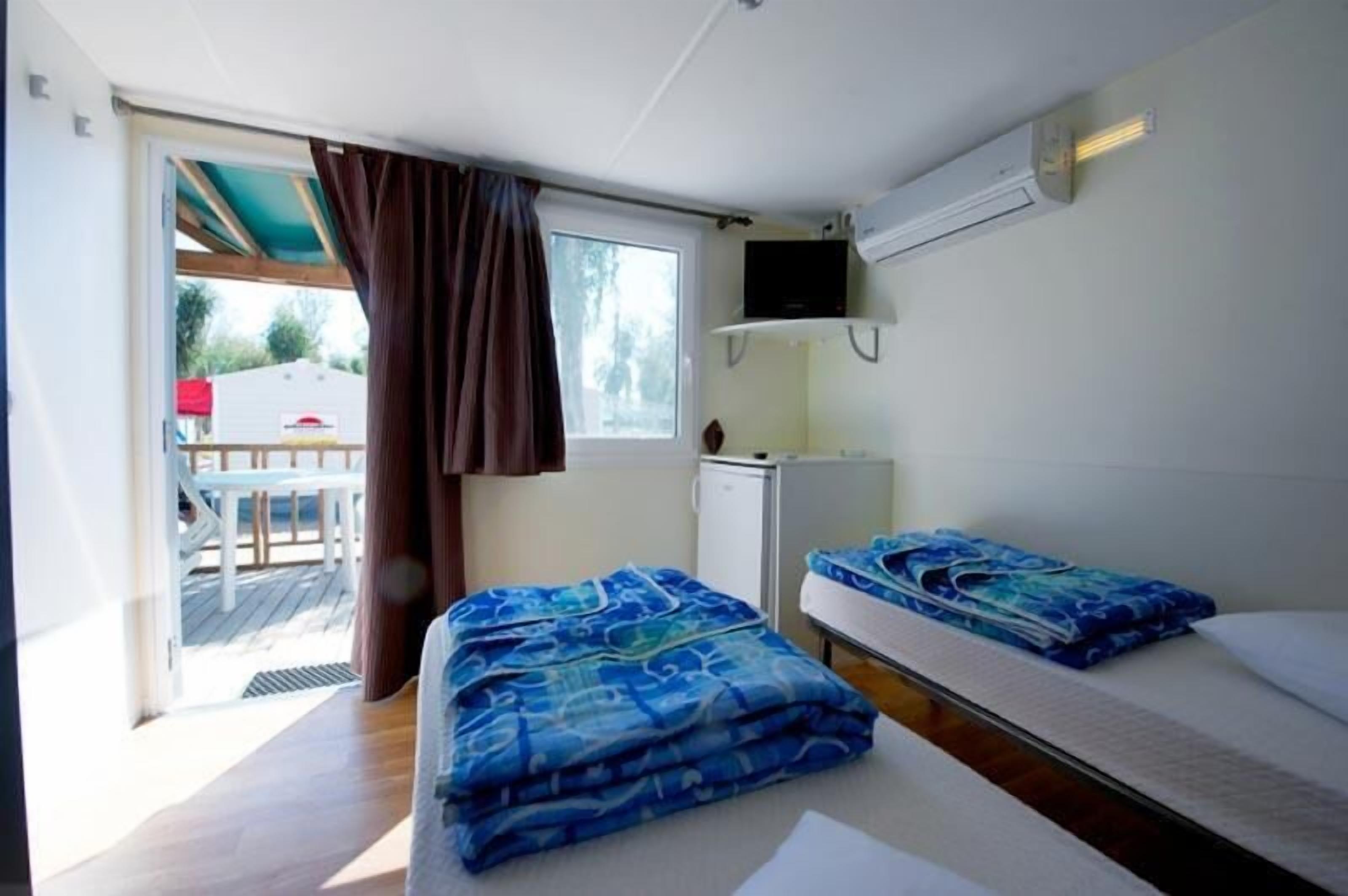 Ein Zimmer mit zwei Betten, einem kleinen Schreibtisch, einem Fenster mit Vorhängen und einem an der Wand befestigten Fernseher.