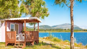 Una cabaña de madera con un porche, una mesa y sillas, con vista a un lago y montañas al fondo.
