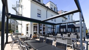 An outdoor patio area with tables and chairs, a glass enclosure, and a building in the background. - Hotel Gulliver (Käerjeng)