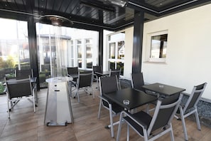 An outdoor patio with a round table and chairs, a modern heater, and large windows offering views of the surrounding area. - Hotel Gulliver (Käerjeng)