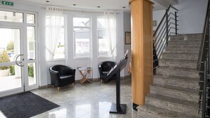 A lobby with a reception desk, two black leather chairs, a small table, a standing art display, and a staircase. - Hotel Gulliver (Käerjeng)