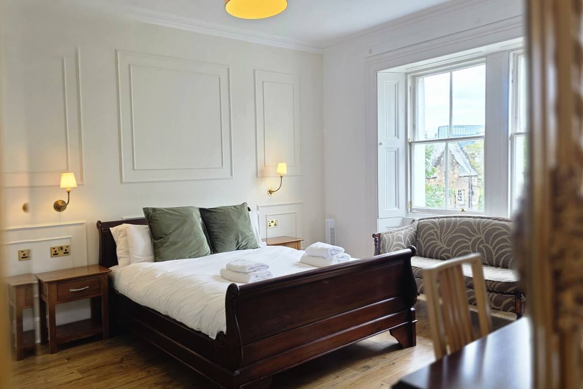 A bedroom with a wooden bed, two bedside lamps, a nightstand, a sofa, and a window with a view.