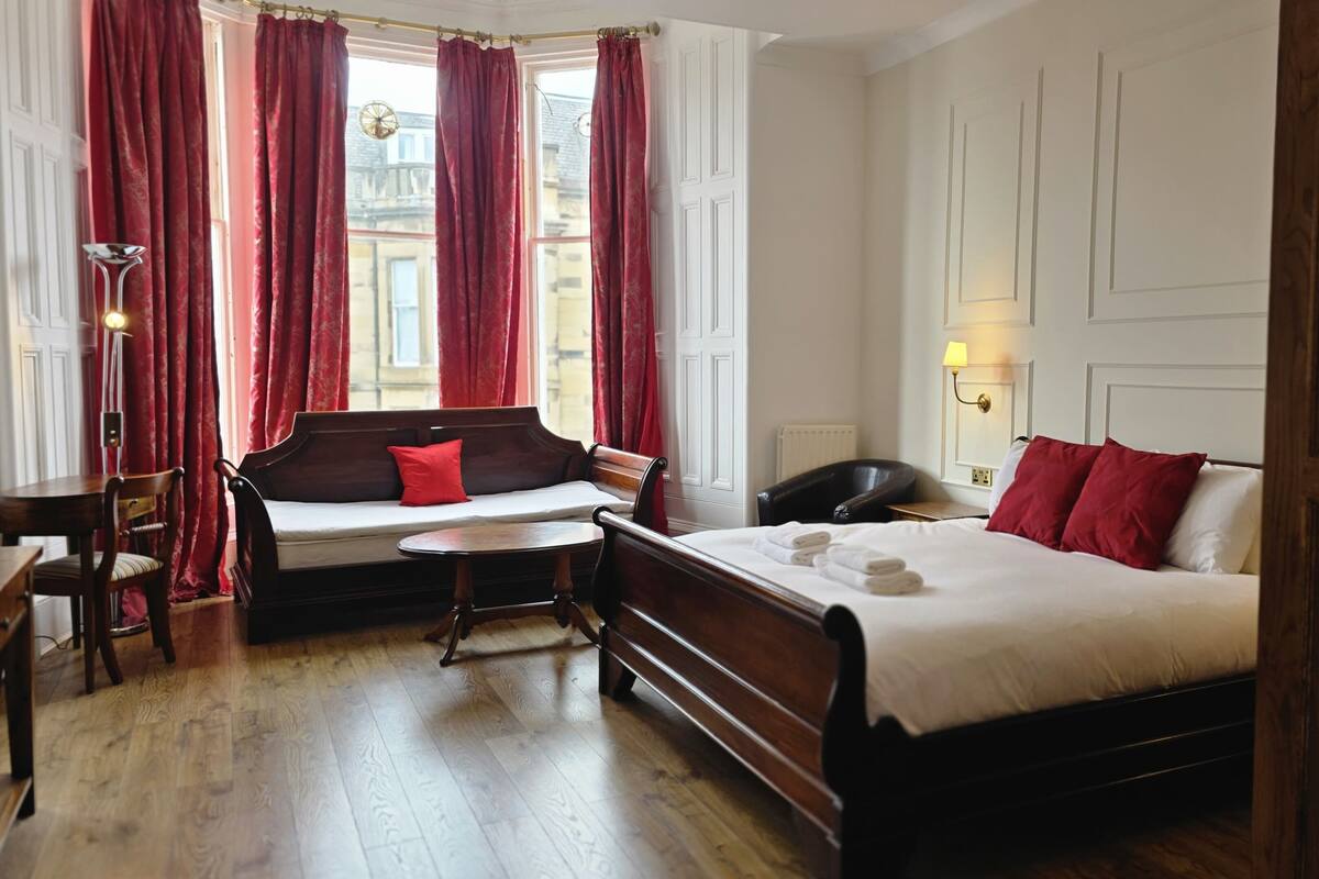 A hotel room with a wooden bed, a bench with red cushions, a small table, and a lamp.