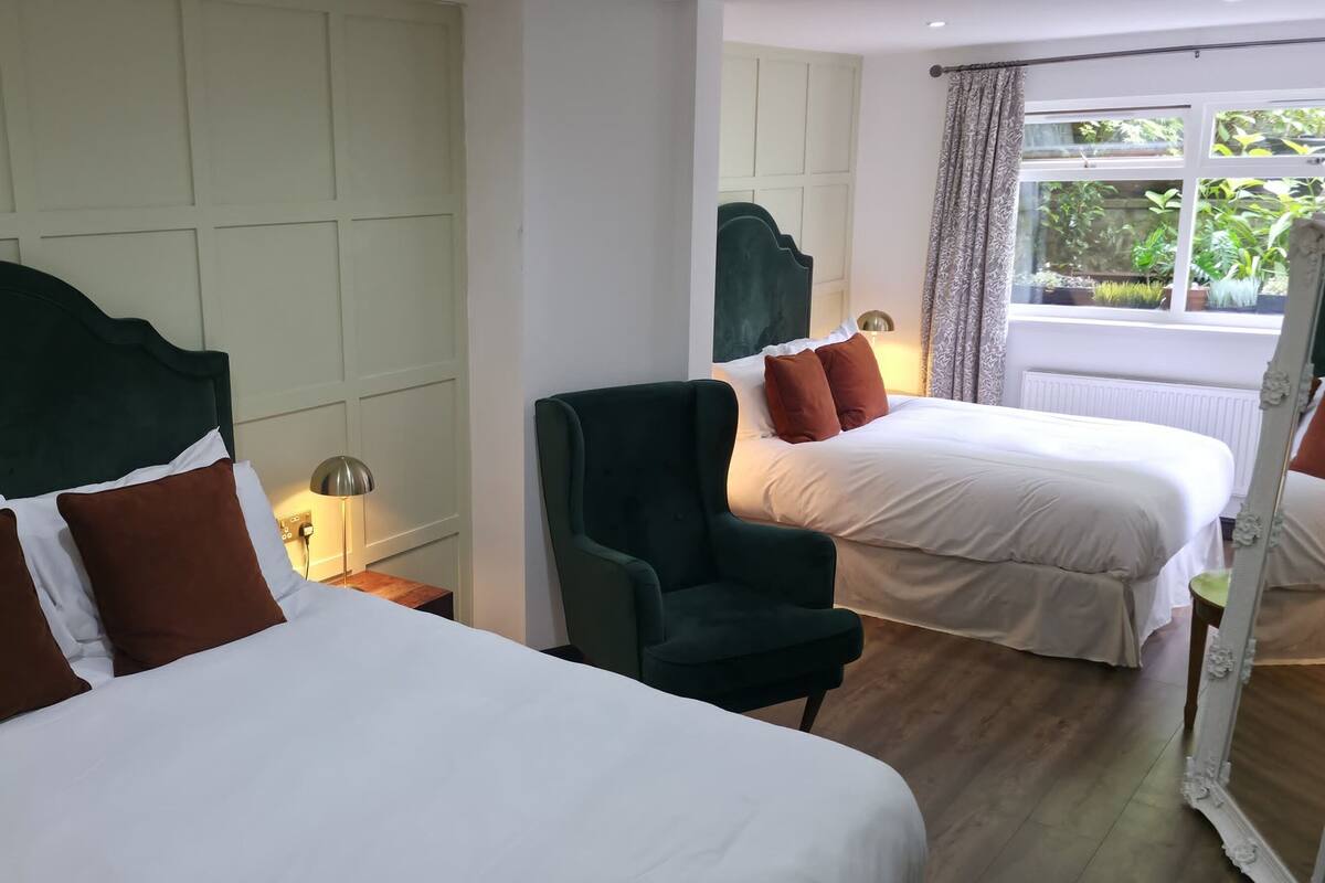 A hotel room with two beds, a chair, a mirror, and a window with a view of greenery.