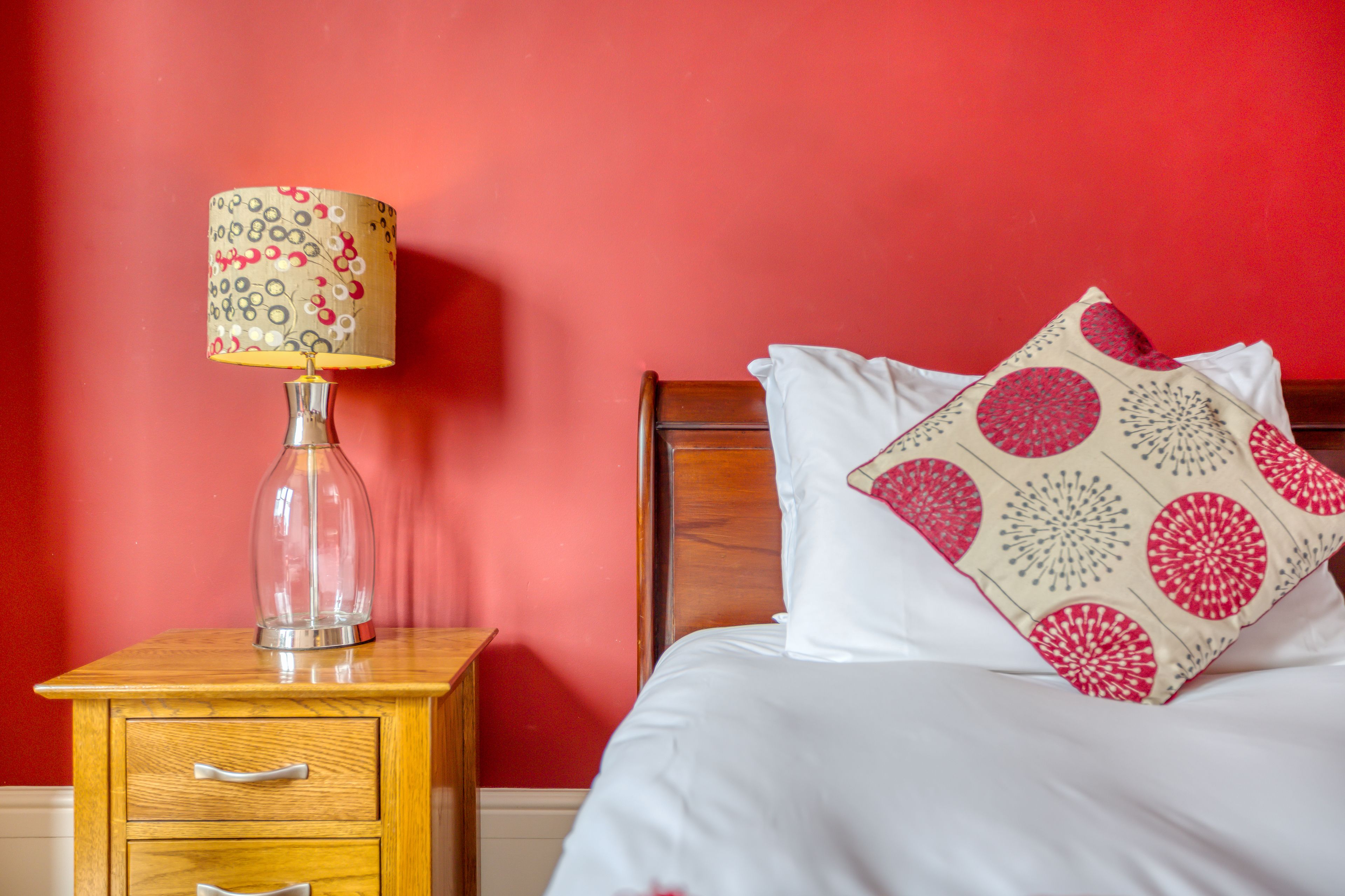 Una cama bien hecha con un almohadón blanco y uno estampado, una mesita de noche de madera con una lámpara, y una pared roja.