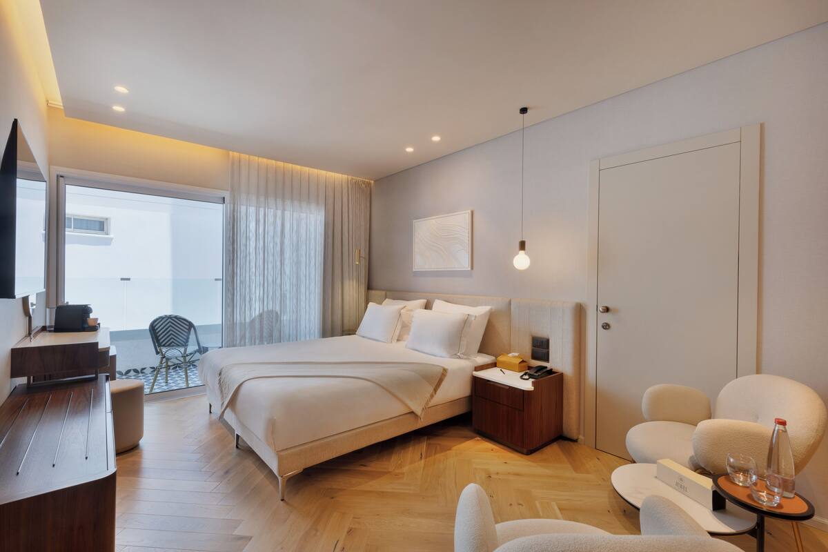 Habitación de hotel moderna con una cama grande, un escritorio, una silla, una mesita con vasos y una botella, y vistas al exterior.