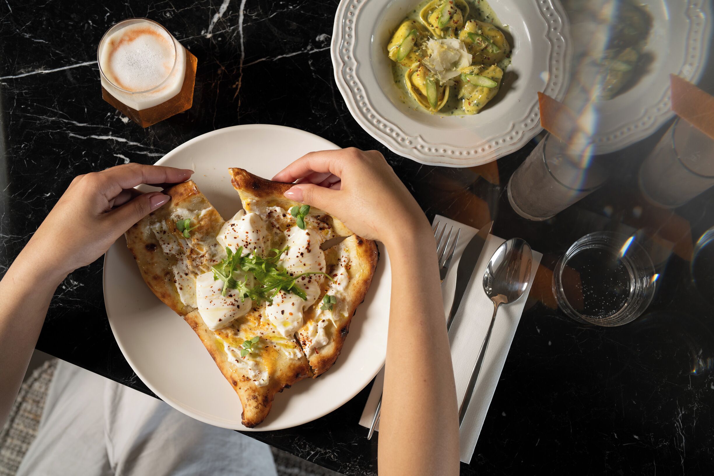Een persoon houdt een bord pizza met toppings vast, samen met een glas koffie, op een marmeren tafel.