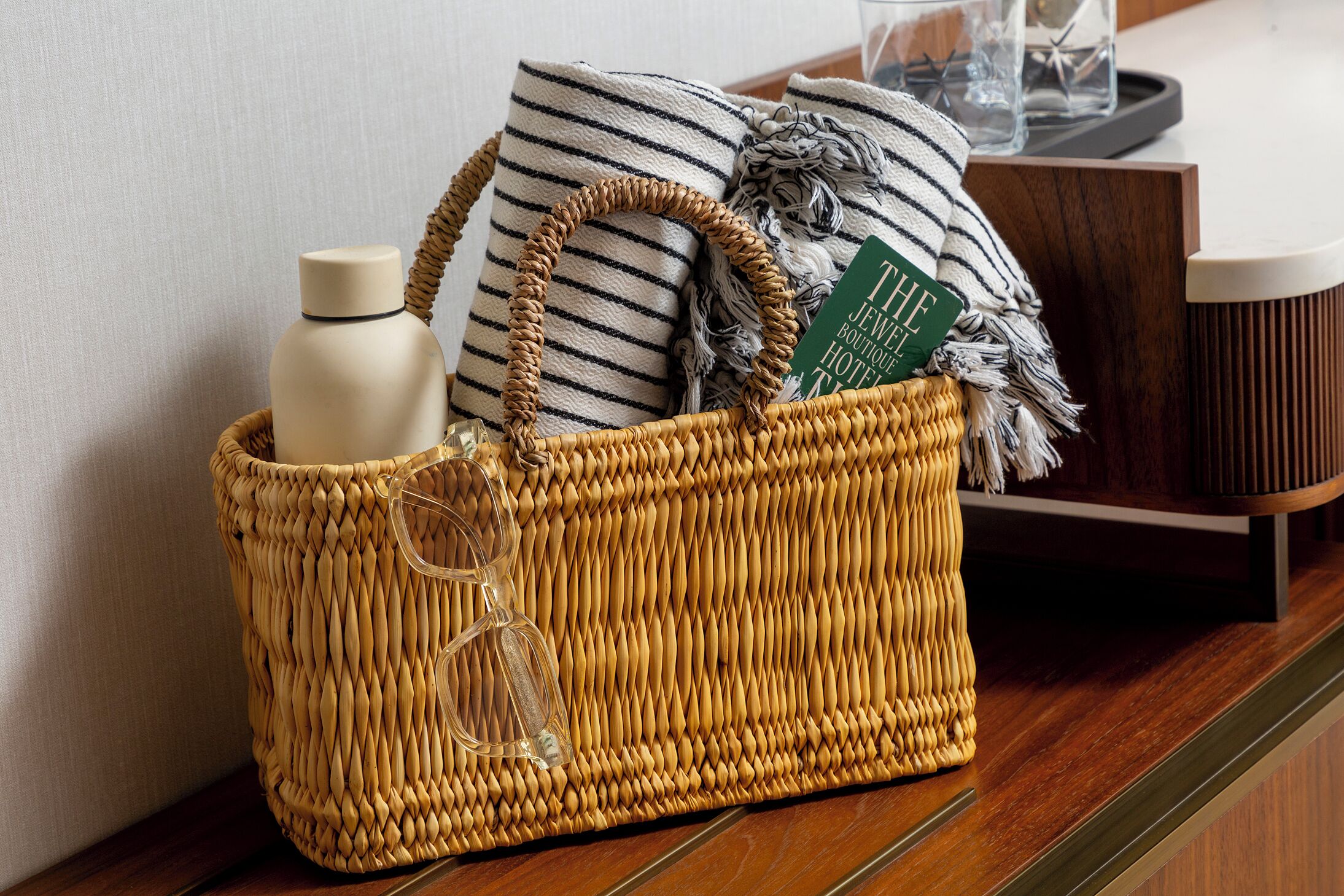 Una cesta de mimbre con una botella, un libro y una almohada a rayas sobre una superficie de madera.