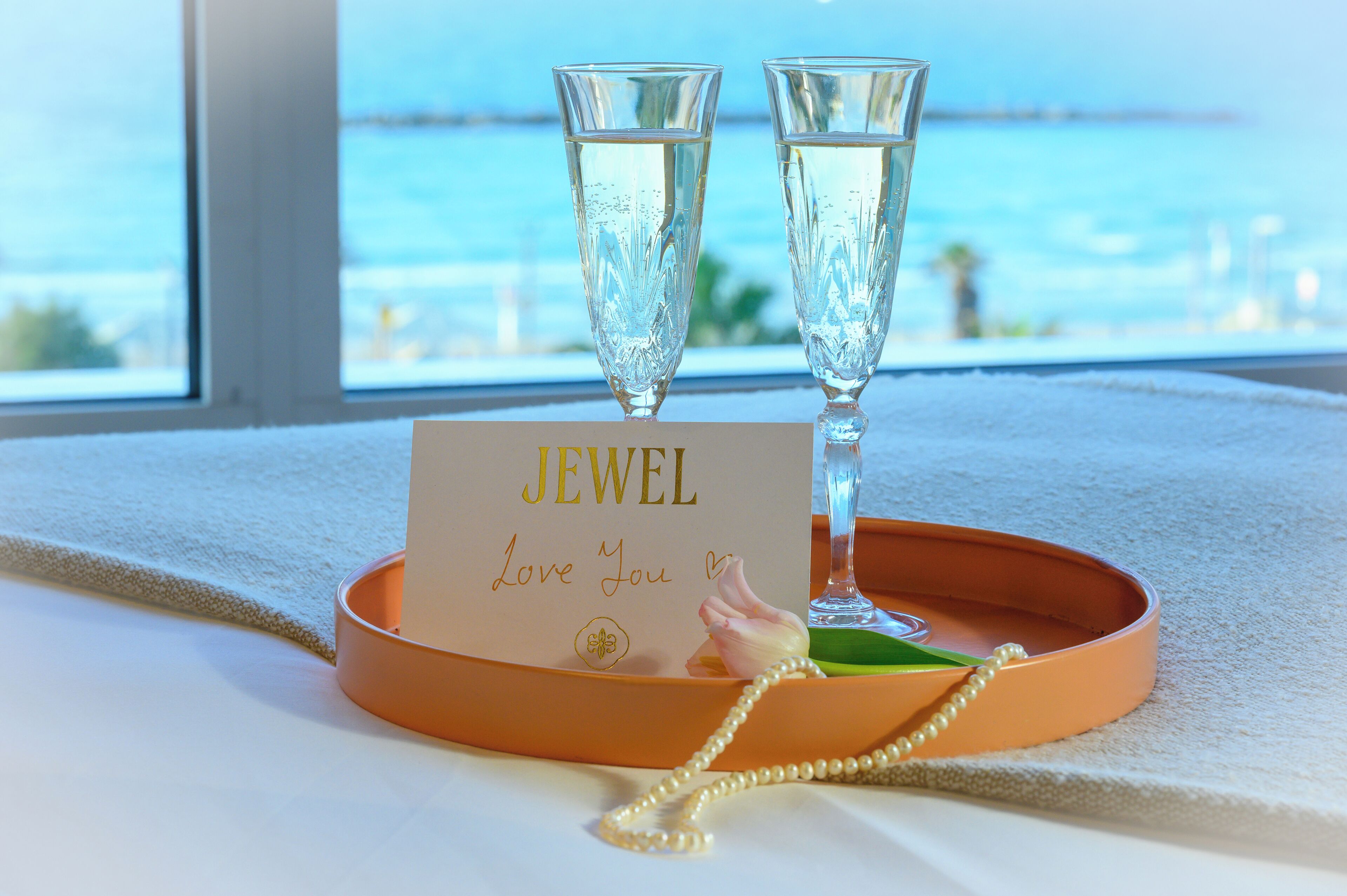 Uma bandeja com duas taças de champanhe e um cartão com a mensagem "JEWEL Love You" sobre uma mesa com vista para o mar.