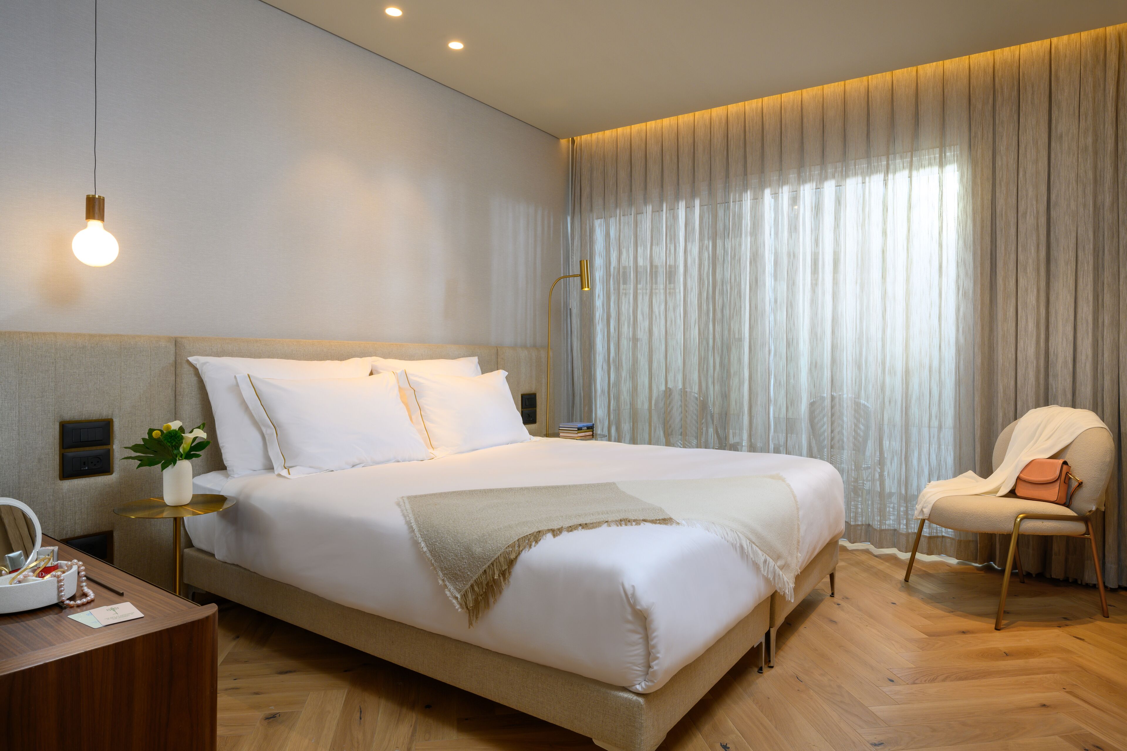 A modern hotel room with a large bed, a bedside table with a lamp, a chair, and a view of the outside through sheer curtains.
