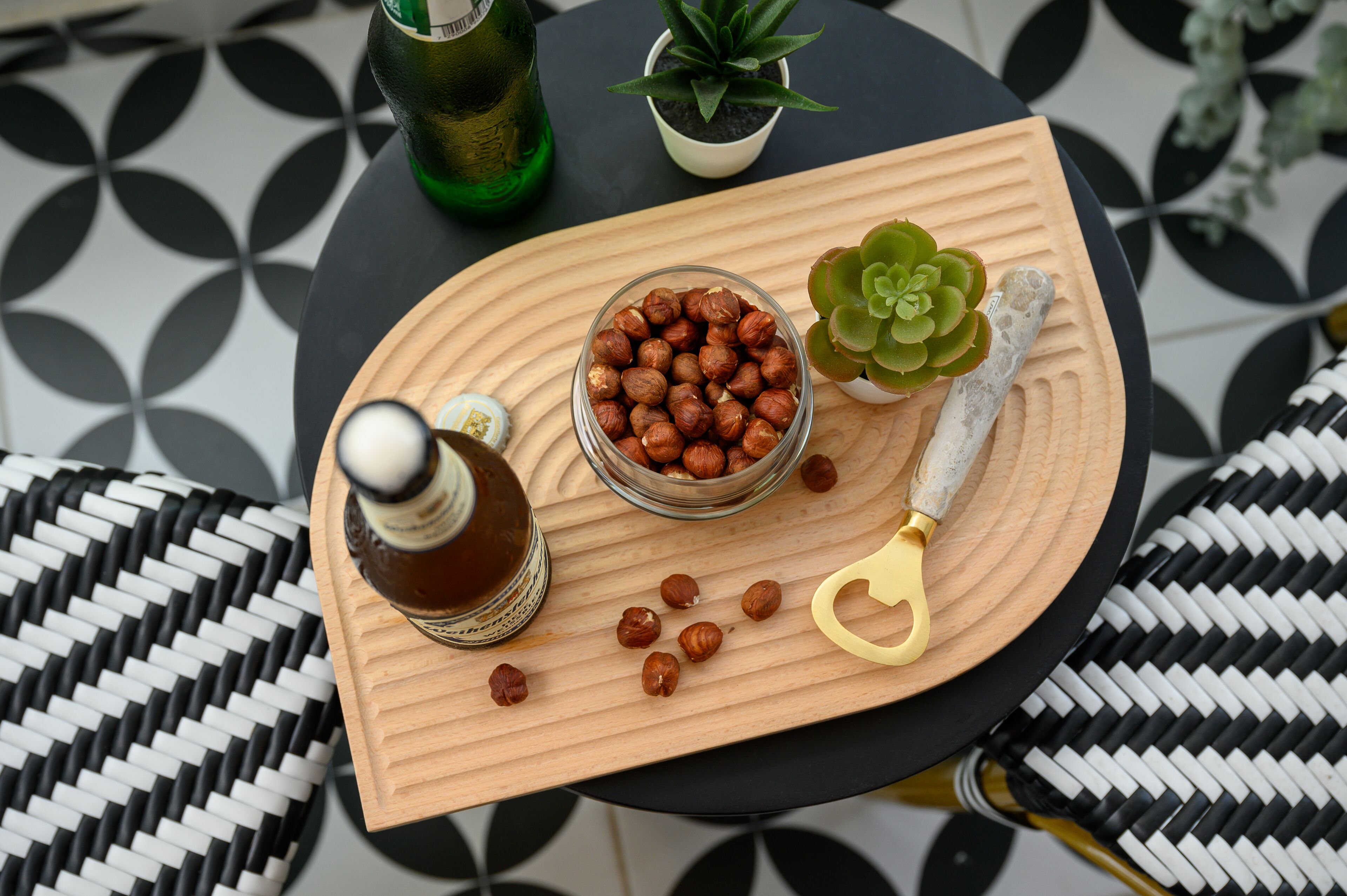 Una bandeja de madera con un tazón de vidrio lleno de nueces, una botella y un cuchillo sobre una mesa con piso de estampado blanco y negro.