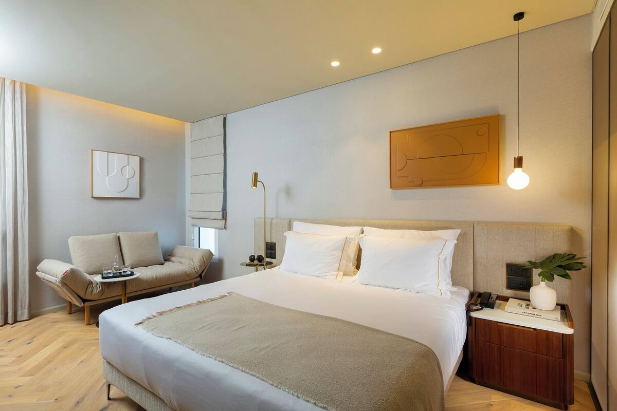 Una habitación de hotel moderna con una cama grande, un sofá, una mesita de noche con lámpara y una obra de arte en la pared.