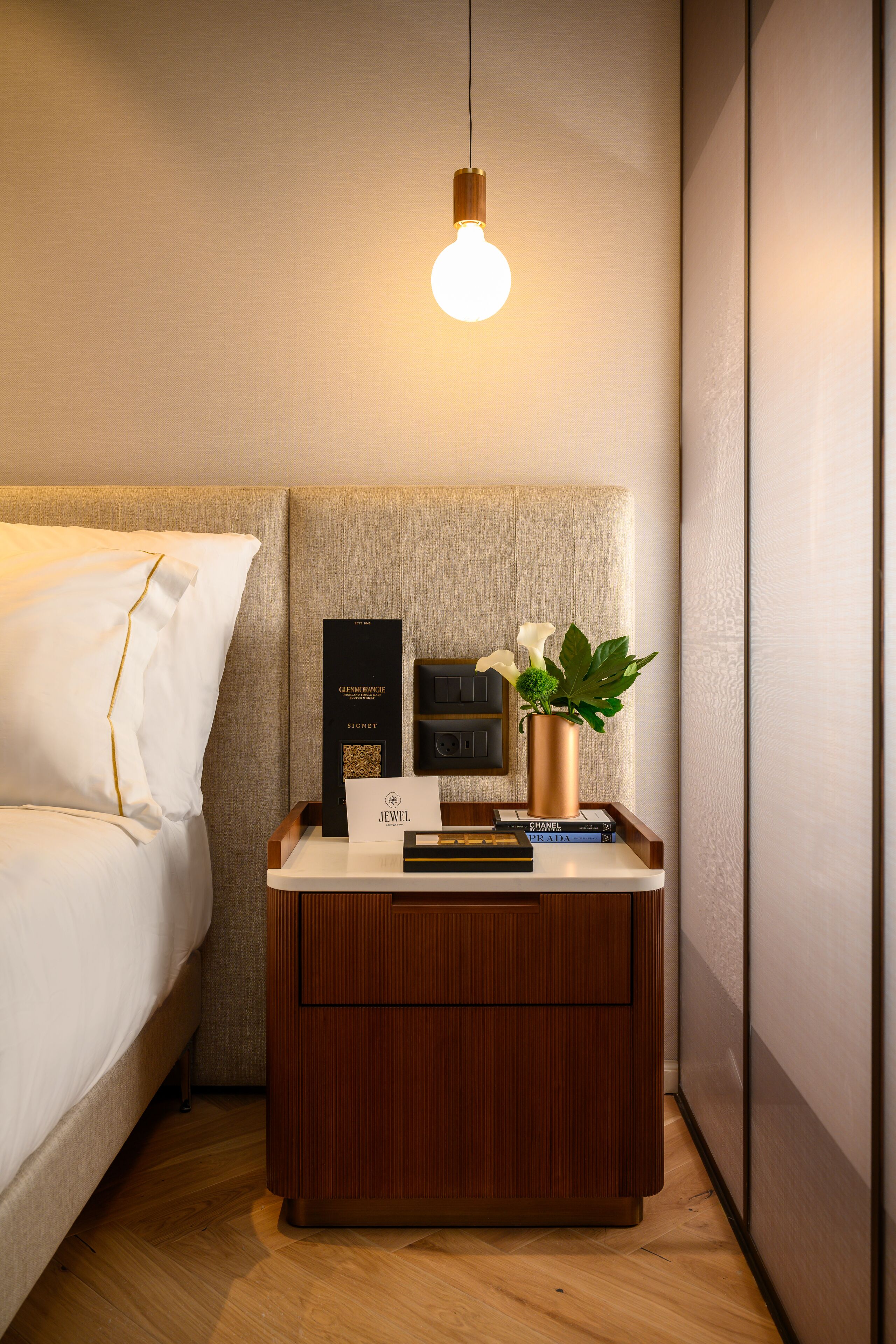 Quarto de hotel bem arrumado com mesa de cabeceira de madeira, cama com roupa de cama branca, luminária pendente e uma pequena planta em vaso.