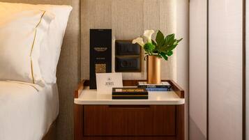 Quarto de hotel bem arrumado com mesa de cabeceira de madeira, cama com roupa de cama branca, luminária pendente e uma pequena planta em vaso.