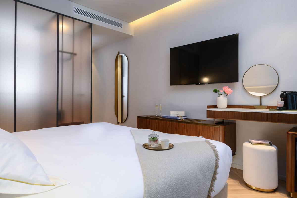 Una habitación de hotel con cama, mesita de noche con bandeja, espejo, televisor de pantalla plana y armario.
