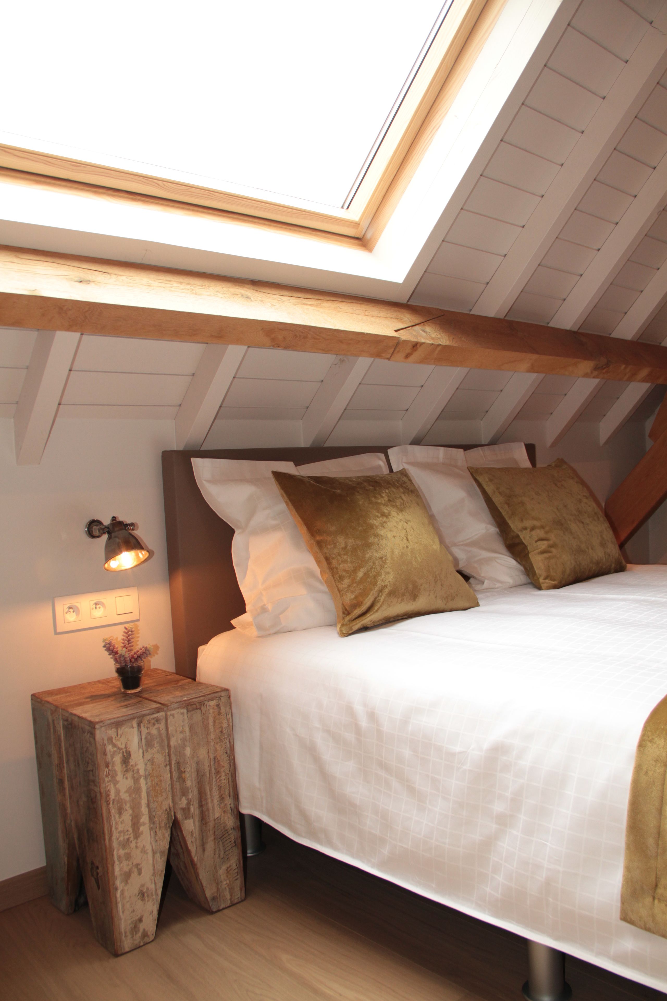 A bedroom with a sloped ceiling, a bed with white bedding and gold pillows, a bedside lamp, and a small wooden nightstand.
