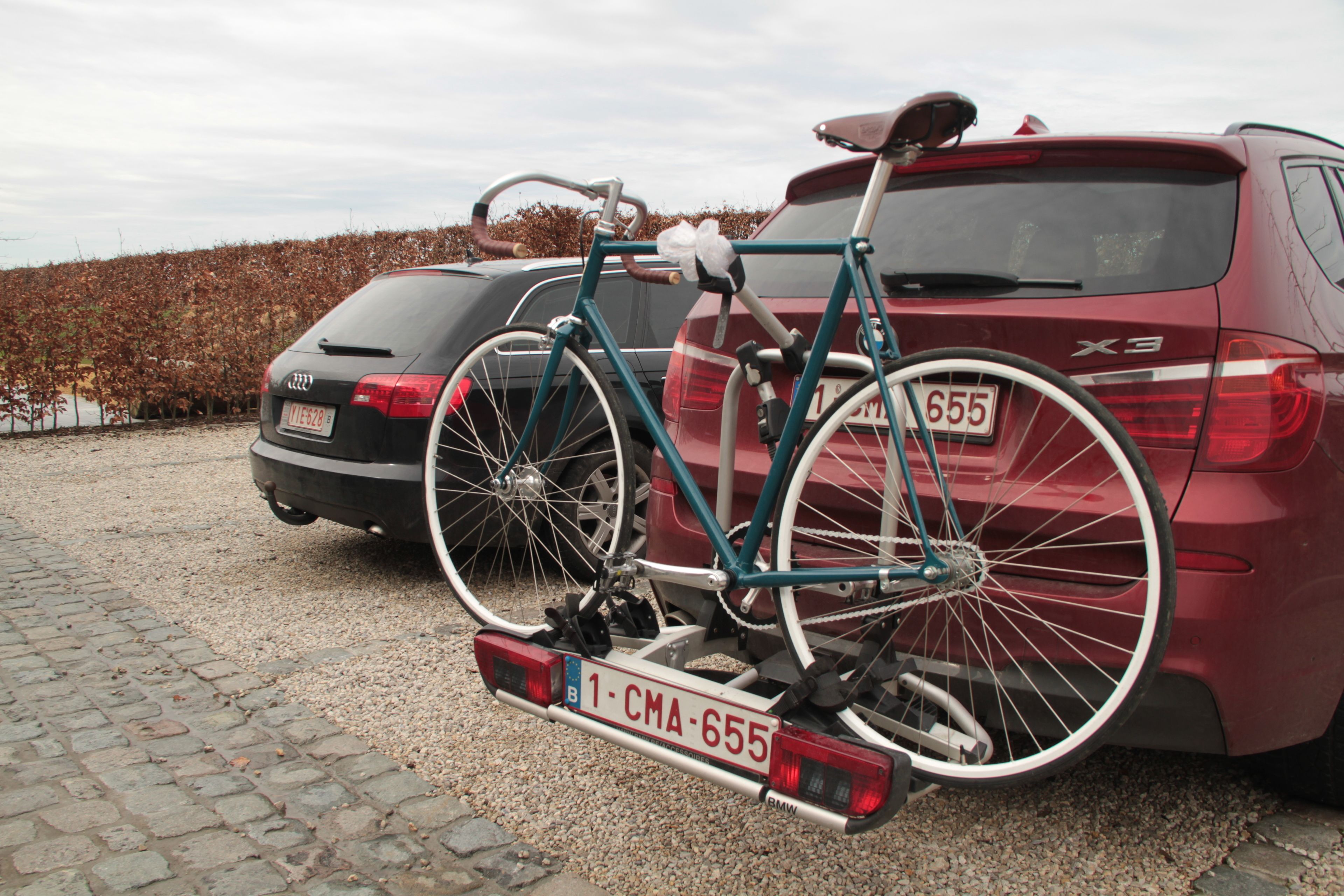 Ein Fahrrad mit einem Nummernschild, befestigt am Heck eines roten Autos, das auf einem Schotterparkplatz steht.