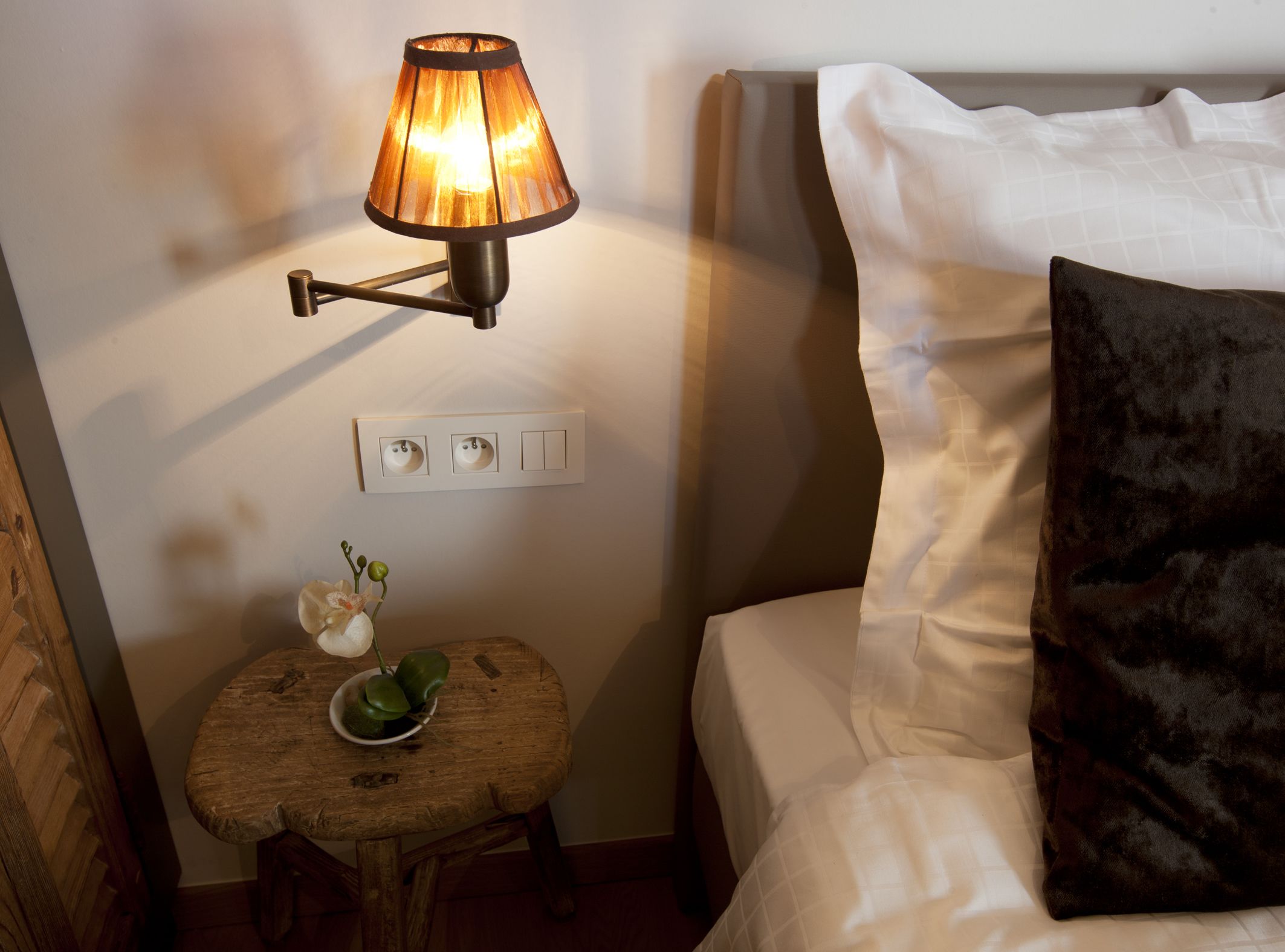 A neatly made bed with a white headboard and a dark brown pillow, a small wooden table with a green plant, and a wall-mounted lamp with a warm light.
