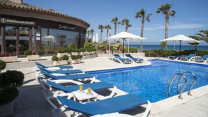 A pool area with lounge chairs, umbrellas, and a building with signage. - Cala Cristal by Pierre & Vacances Premium (Mont-roig del Camp)