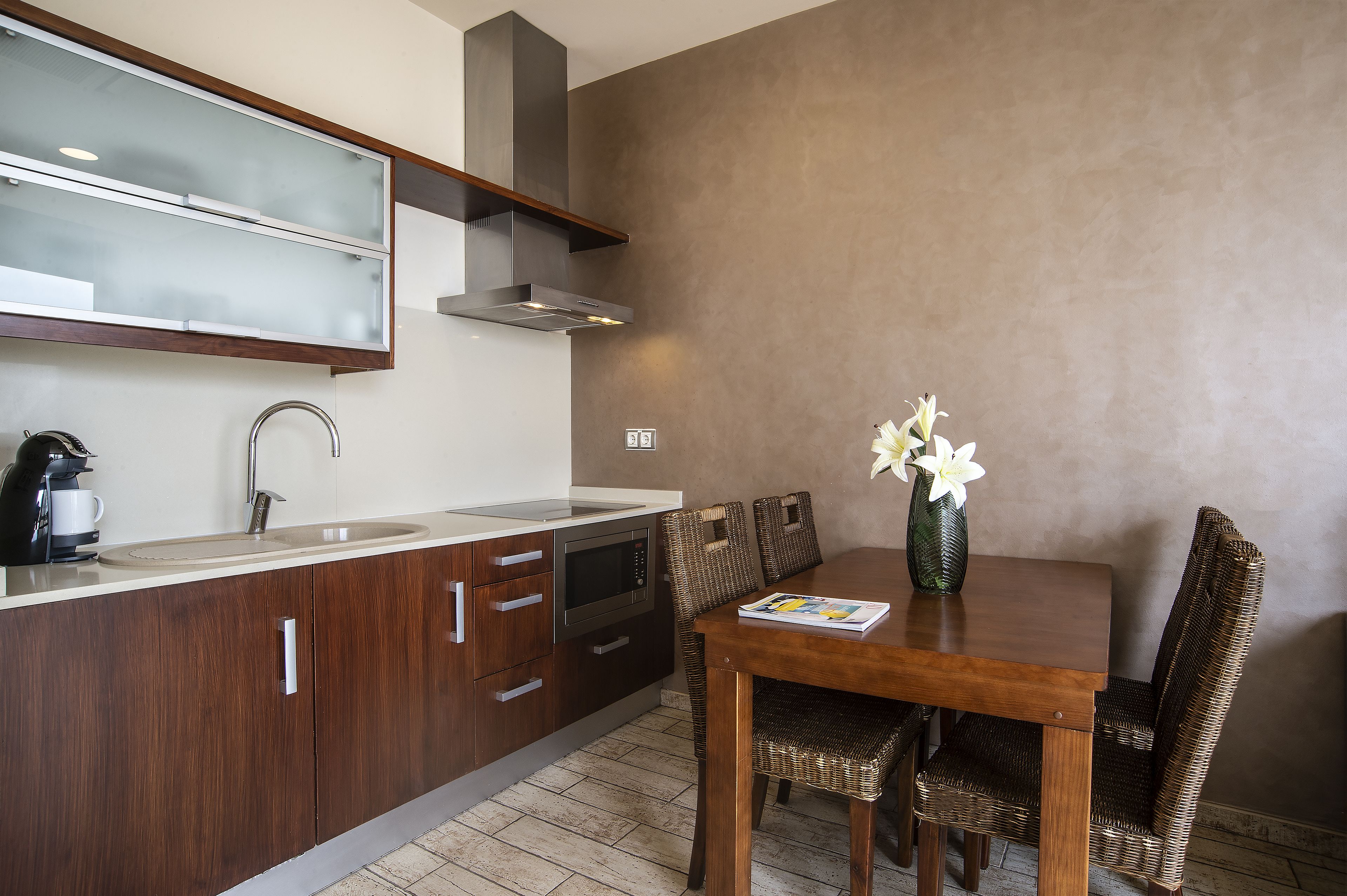 A modern kitchen with a built-in oven, microwave, and stainless steel sink. There is a dining table with wicker chairs and a vase with flowers.