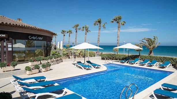 A pool area with lounge chairs and umbrellas, a building with a sign, and palm trees. - Cala Cristal by Pierre & Vacances Premium (Mont-roig del Camp)
