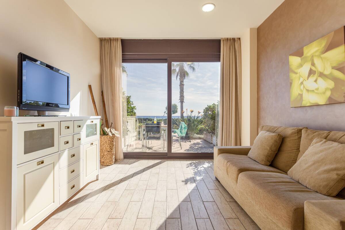 A modern living room with a flat-screen TV, a beige sofa, and a large sliding glass door leading to a balcony with a view of palm trees and the sea.