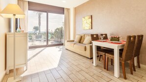 A modern living room with a sofa, dining table, and chairs. There is a large window with a view of palm trees and a balcony. - Cala Cristal by Pierre & Vacances Premium (Mont-roig del Camp)
