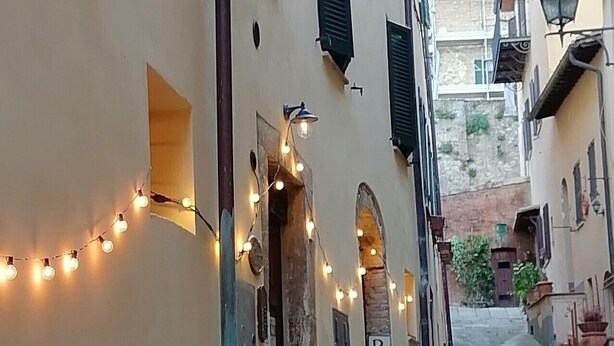 Une ruelle étroite bordée de bâtiments des deux côtés, avec des guirlandes lumineuses suspendues au-dessus et une enseigne indiquant « CHAMBRES ».