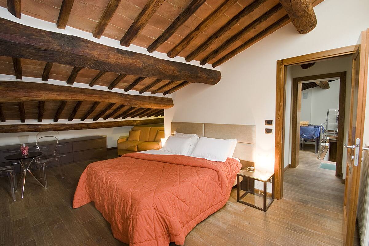 Une chambre avec un plafond en bois, un lit recouvert d’une couverture orange, un bureau avec une chaise et une petite table avec une lampe.
